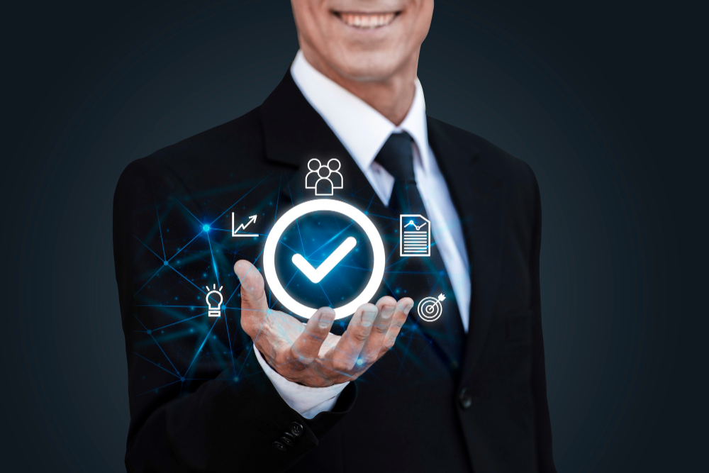 The image shows a businessman in a suit and tie, smiling and holding out his hand. Above his open hand, there is a glowing circular icon with a checkmark in the center, symbolizing approval or completion. Surrounding the checkmark are various digital icons representing business-related concepts such as graphs, documents, people, and targets. The background is dark, which highlights the glowing icons and adds a futuristic, high-tech feel to the image. The overall impression is one of confidence, professionalism, and successful management.