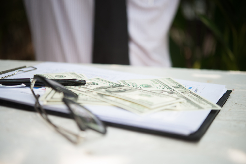 A close-up photo of a table with scattered US dollar bills, a pair of eyeglasses, and a pen resting on top of a document or clipboard. The background is slightly blurred, with a person standing and some greenery visible, suggesting an outdoor setting. The scene conveys a sense of financial or business activity, such as a transaction, signing documents, or reviewing financial information.