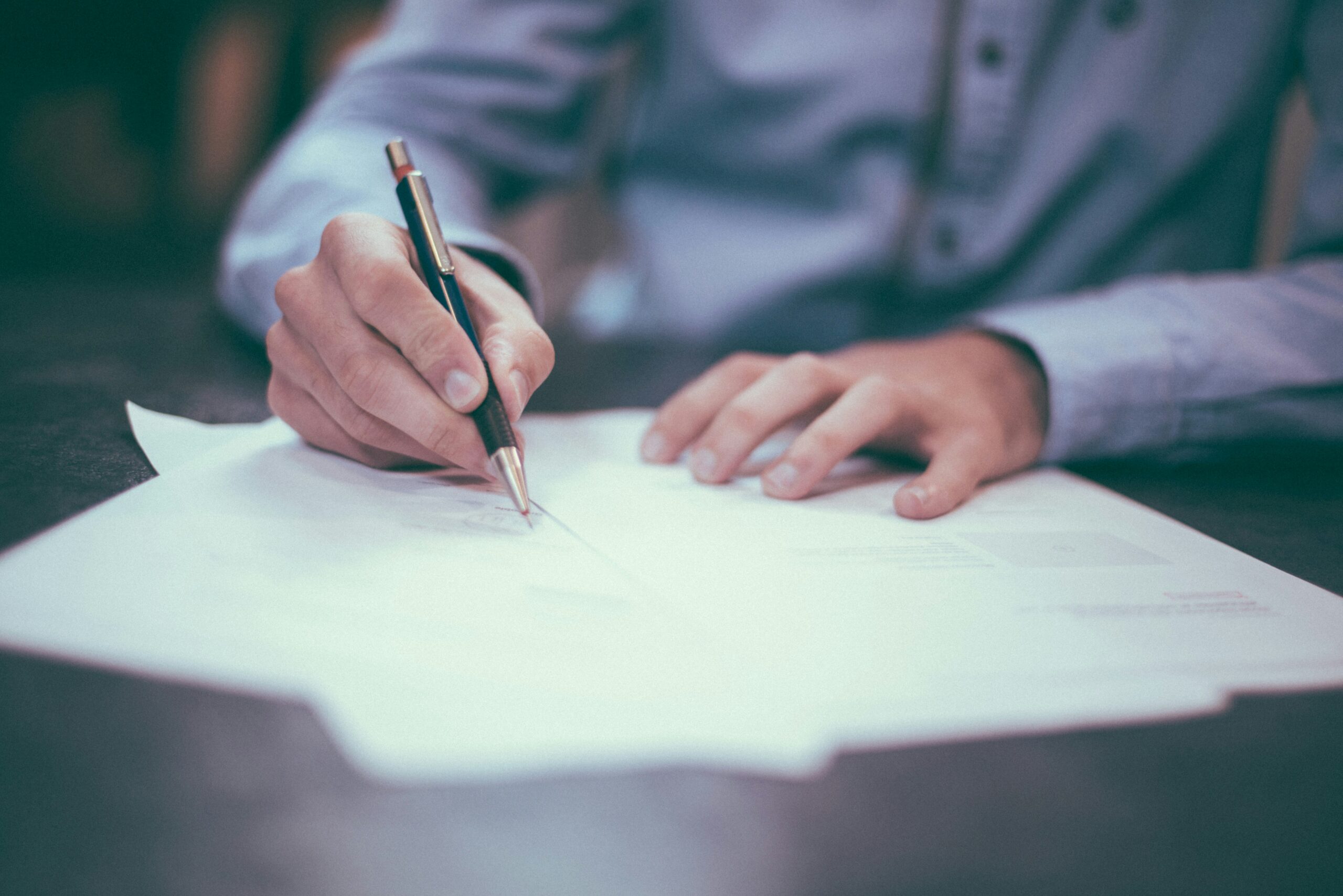 A close-up photo of a person signing documents. The individual is wearing a light blue shirt and is holding a pen, poised to write on one of several papers spread out on a dark wooden table. The focus is on the hands and the documents, emphasizing the act of signing or reviewing important paperwork. The background is blurred, keeping attention on the task at hand, suggesting a professional or business setting.