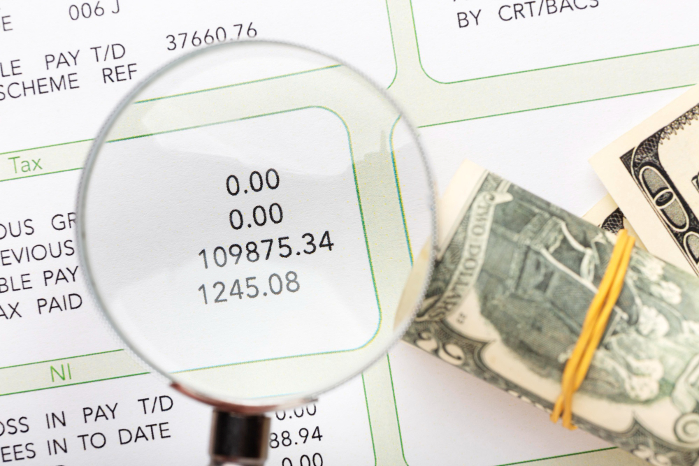 A magnifying glass focusing on a detailed financial document displaying various numbers, with a rolled stack of dollar bills secured by a rubber band nearby. The scene emphasizes careful examination and analysis of financial data, suggesting activities related to auditing, budgeting, or financial scrutiny.