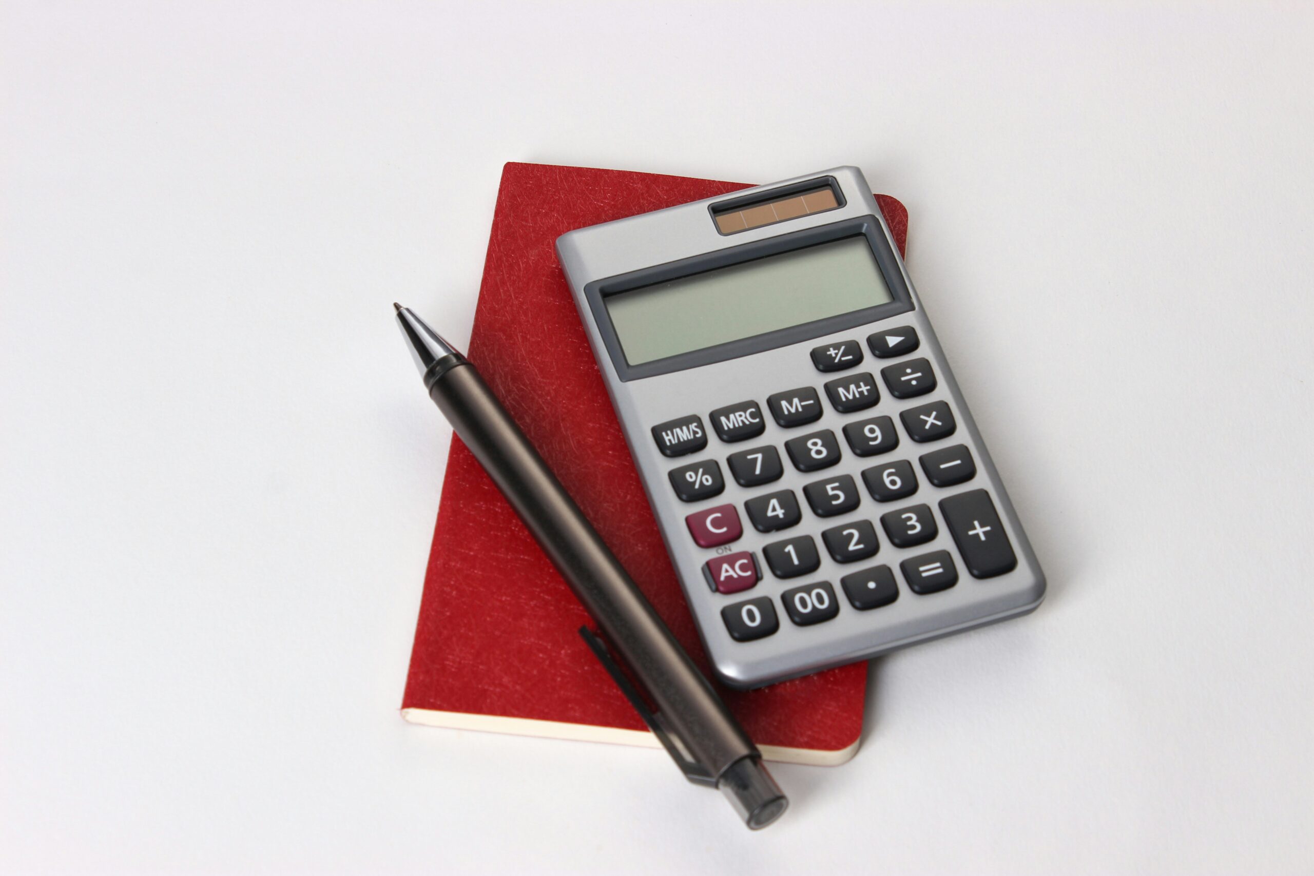 A simple arrangement of a calculator, a black pen, and a small red notebook placed on a clean white surface. The scene suggests a minimalistic and organized setup for basic calculations and note-taking.