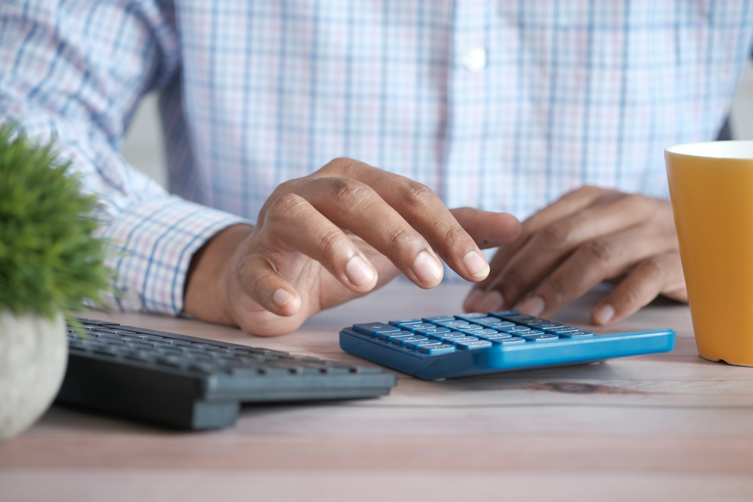 A close-up of a person using a calculator at a desk. The individual is wearing a checkered shirt, and their hand is pressing buttons on the calculator. Nearby, there is a keyboard, a small potted plant, and a yellow coffee mug. The scene suggests a focused work environment, likely involving financial calculations or data entry.