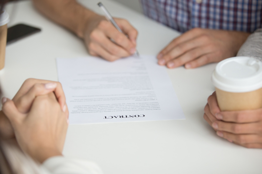A photo of three people sitting at a table, focused on a contract document. One person is signing the contract with a pen, while another holds a coffee cup, and the third person has their hands clasped together. The scene suggests a business meeting or agreement being finalized. The table is white, and the atmosphere appears professional and collaborative. The contract is clearly labeled "CONTRACT" at the top, emphasizing the formal nature of the document.