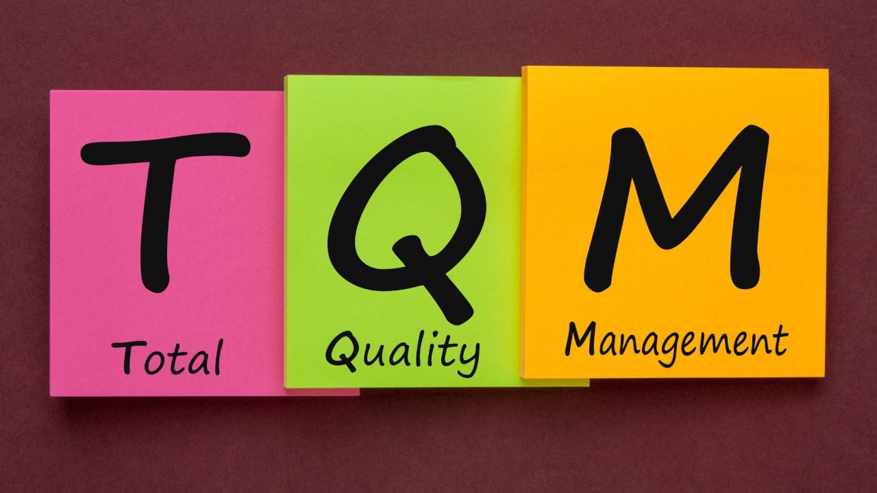 A colorful graphic with three sticky notes arranged side by side. The notes display the acronym "TQM," which stands for Total Quality Management. The "T" is on a pink note labeled "Total," the "Q" is on a green note labeled "Quality," and the "M" is on an orange note labeled "Management," all set against a dark background.