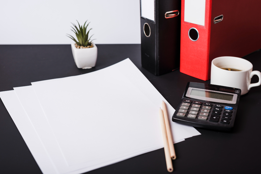 A neatly organized desk with blank sheets of paper, two pencils, a small potted plant, a calculator, a cup of coffee, and two binders (one red and one black). The scene suggests a clean and prepared workspace, ready for work, planning, or organization tasks.