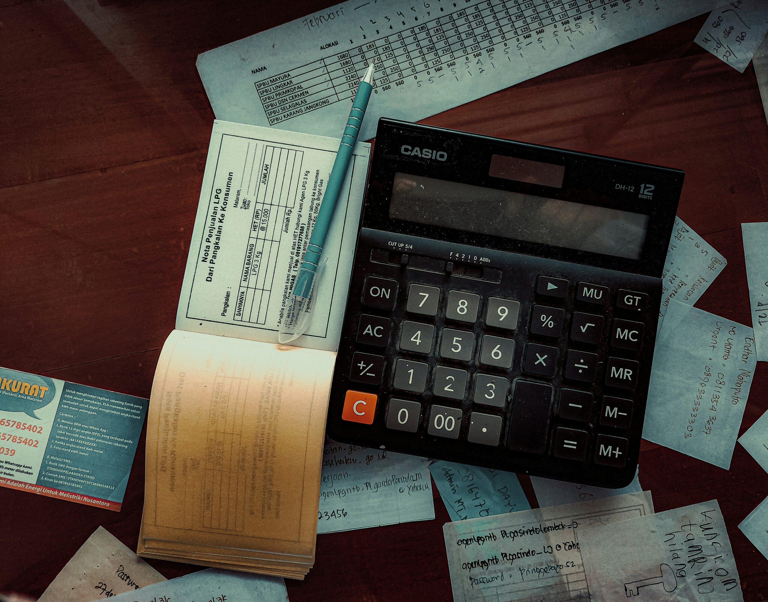 A top view of a desk cluttered with financial documents, receipts, and a large calculator. A pen rests on an open ledger, suggesting accounting or budgeting activities. The scene conveys a sense of meticulous financial management and record-keeping.