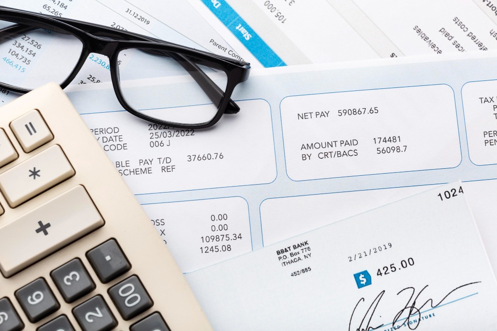 A pair of black-framed glasses rests on top of several documents, including invoices and a bank statement. A calculator with large buttons is positioned to the left, indicating financial calculations or accounting work in progress. The documents display various financial figures, such as net pay, tax, and amount paid, providing a clear context of financial management and record-keeping.