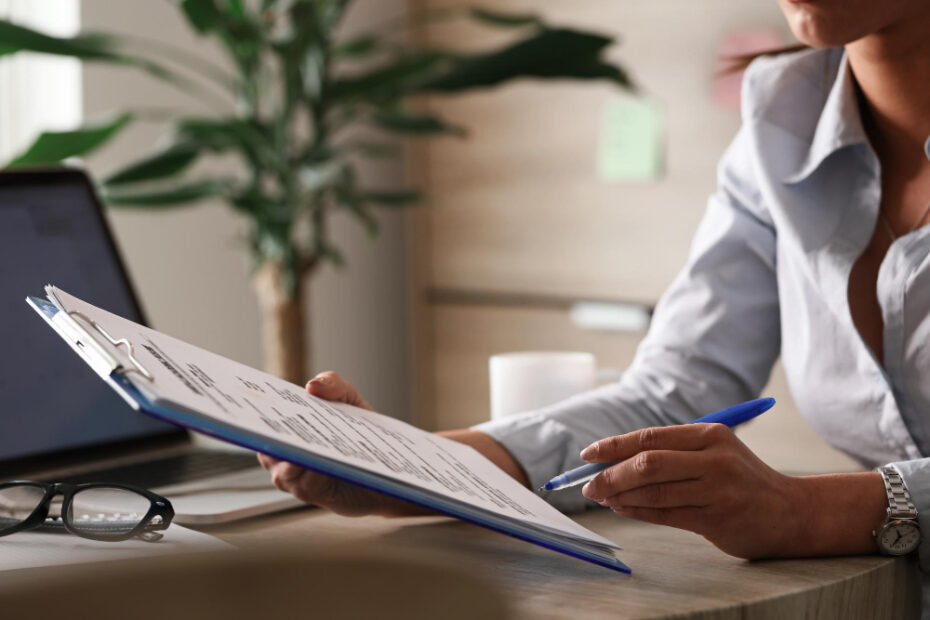 A person is sitting at a desk holding a clipboard with documents. The individual is reviewing the papers with a pen in hand, indicating they might be making notes or filling out forms. A pair of glasses and an open laptop are on the desk, suggesting a work environment. In the background, there is a blurred view of a plant, adding a touch of greenery to the workspace.