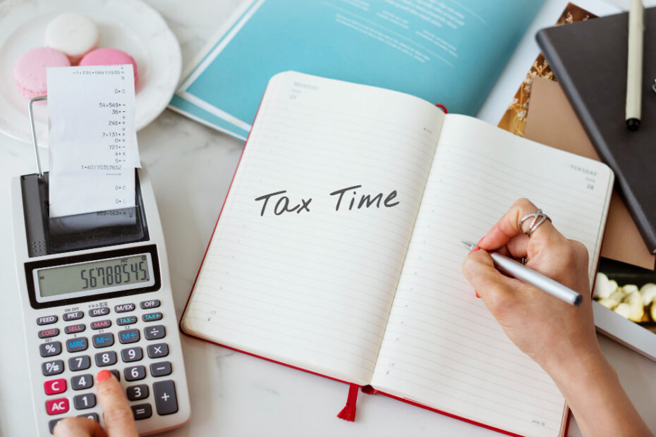 The image shows a person working on their taxes. They are using a calculator that displays numbers and a receipt is visible emerging from the top of the calculator. The person's left hand is pressing buttons on the calculator, while their right hand is writing in a notebook with the words "Tax Time" written at the top of the page. The scene is set on a desk with various items including a book, a pen, and some pink macarons in the background. The overall atmosphere suggests a focus on financial planning or tax preparation.