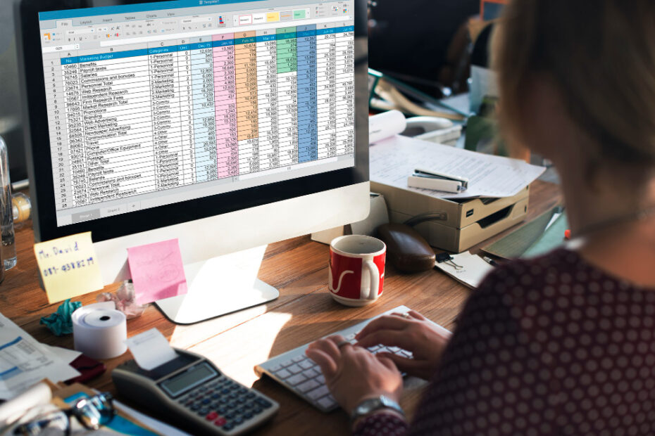 A person is sitting at a desk, working on a computer with a large monitor displaying a detailed spreadsheet. The spreadsheet contains multiple rows and columns with data organized by categories, such as "Benefits," "Payroll," and "Marketing," each marked with different colors. The workspace is cluttered with office supplies, including a calculator, sticky notes, a coffee mug, and various papers. The setting suggests a busy, organized environment where financial or data management tasks are being performed.