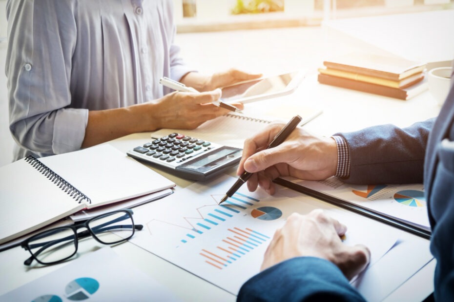 Two professionals are working together at a desk filled with financial documents, graphs, and charts. One person is using a calculator while the other is pointing to a chart with a pen, indicating a collaborative discussion on financial data. There are notebooks, a tablet, and eyeglasses on the desk, suggesting a focus on accounting or financial analysis. The setting is bright, with natural light illuminating the workspace.