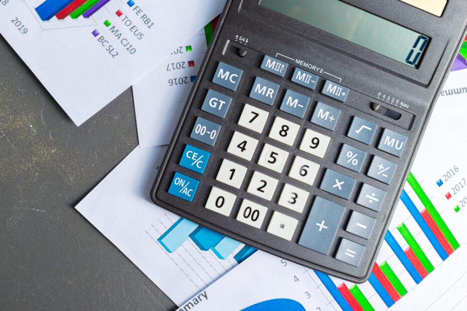 A large calculator is placed on top of several colorful financial charts and graphs, displaying various data trends. The charts include bar graphs with data from different years, showcasing financial or business performance metrics. The calculator's screen is currently blank, indicating it's ready for use. The image conveys a focus on financial analysis, budgeting, or accounting tasks.