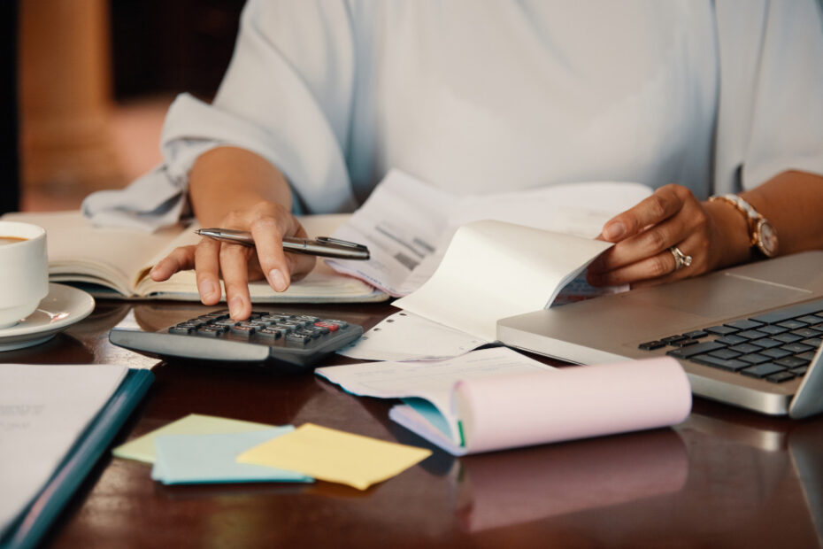 A person is sitting at a desk working with financial documents. They are using a calculator with one hand while holding a pen in the other. There are papers, a laptop, and sticky notes on the desk, indicating they are engaged in accounting or bookkeeping tasks. The scene conveys an image of focused financial management or office work.