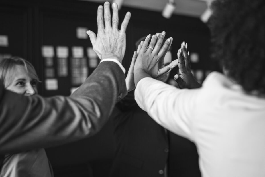 A black and white image showing a diverse group of professionals in an office environment, all reaching in for a collective high-five. The team members are smiling, displaying camaraderie and unity. This gesture symbolizes teamwork, mutual support, and shared success. The background is blurred, drawing focus to the hands and the expressions of the team members, emphasizing the positive and collaborative atmosphere.