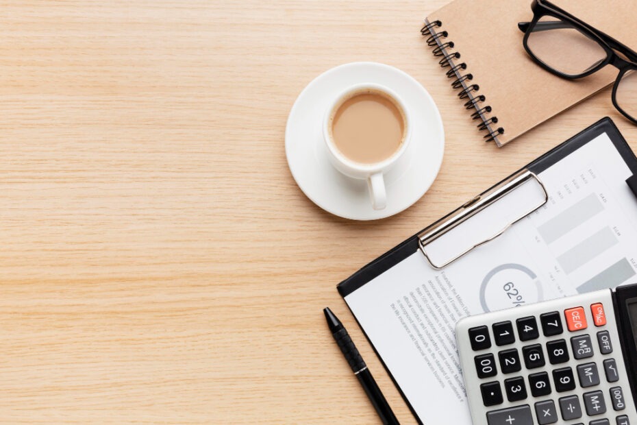 A wooden desk with a coffee cup, a calculator, a clipboard holding a financial report, a black pen, a pair of glasses, and a small spiral notebook. The image portrays a workspace setup for financial or accounting tasks, emphasizing organization and focus on analyzing data.