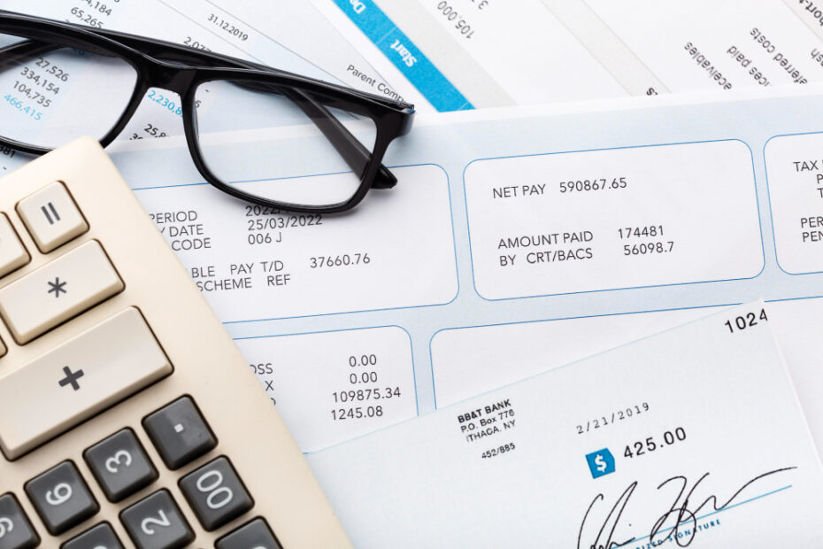 A close-up image of financial documents, including pay slips and a check, alongside a calculator and a pair of eyeglasses. The documents show detailed payment information, such as net pay, tax details, and amounts paid. The eyeglasses are resting on top of the papers, and the calculator is positioned nearby, indicating financial calculations or accounting work.