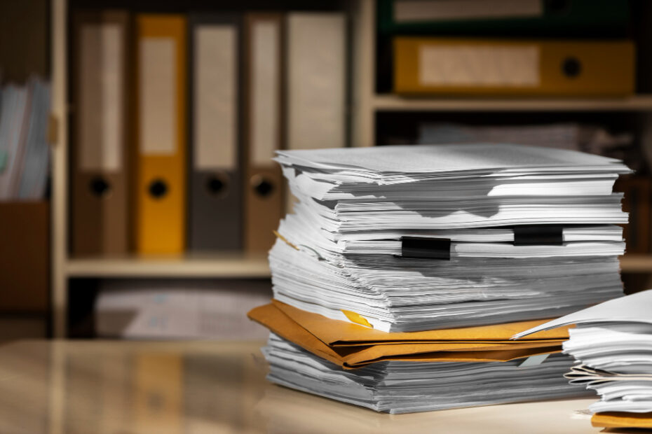 A large stack of paper documents and folders is piled up on a desk, suggesting an overwhelming amount of paperwork. In the background, shelves filled with organized binders and files indicate a traditional, paper-based filing system. The image highlights the contrast between physical document management and the potential benefits of going digital with electronic records or invoices.