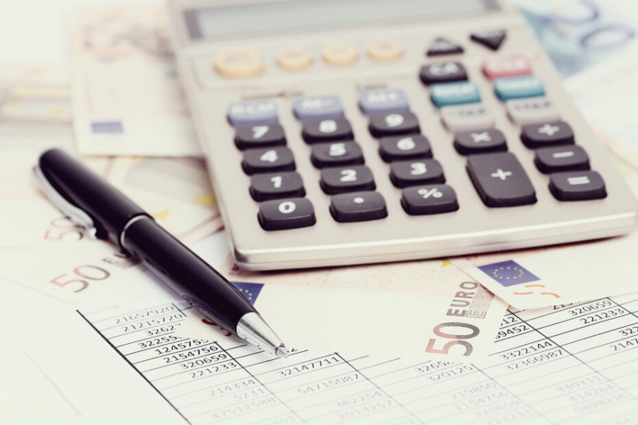 A close-up image of a calculator, a pen, and financial documents placed on a desk, surrounded by several 50 Euro banknotes. The scene suggests financial calculations, budgeting, or tax preparation, with the calculator and pen indicating an active process of working with numbers, possibly related to accounting, tax filing, or financial planning.