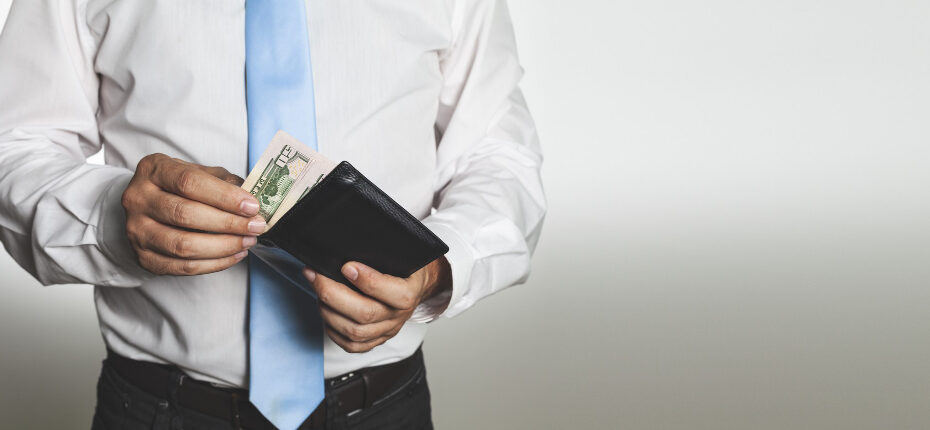 A man wearing a white dress shirt and a light blue tie is holding a black wallet with both hands. He is placing or removing a dollar bill from the wallet. The background is plain and light-colored, drawing attention to the man and the wallet. The image suggests themes related to personal finance, salary, or budgeting.