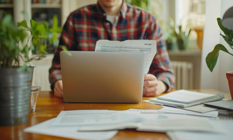 A person in a plaid shirt is working on a laptop at a wooden desk in a well-lit room. The desk is scattered with papers, notebooks, and a pen, indicating a busy work environment. There are several potted plants on the desk and in the background, adding a touch of greenery to the space. The person is holding a document and appears to be focused on their work.