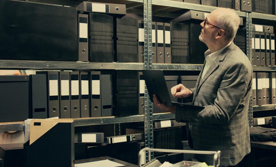 The image shows an older man wearing glasses and a plaid suit, standing in a storage room filled with shelves of black binders and boxes. He is holding a laptop in one hand while looking up at the shelves, seemingly inspecting or searching for something. The setting suggests an archival or records management environment, where physical documents and files are stored and organized systematically. The man's thoughtful expression indicates he may be cross-referencing digital records with the physical files.