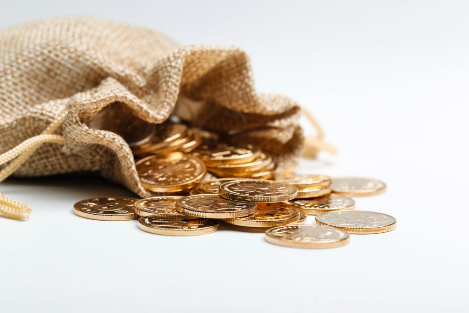 A burlap sack is tipped over on a white surface, spilling out several gold coins. The coins are scattered around the sack, creating a visual impression of wealth or savings. The image suggests concepts like money, savings, wealth, or financial planning.