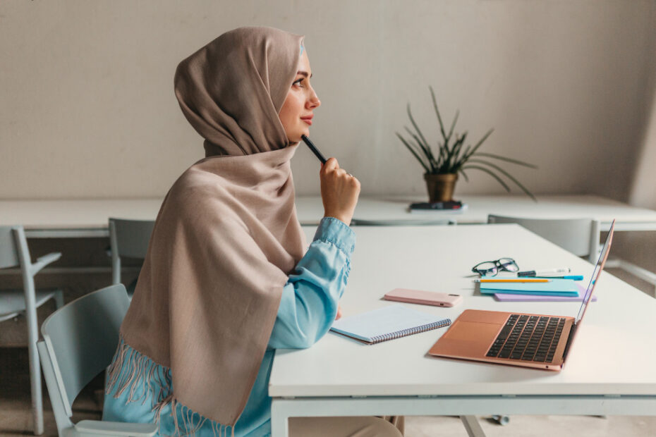 A young woman wearing a beige hijab and a light blue shirt is sitting at a desk with a laptop, notebooks, and pens. She appears to be in deep thought, holding a pen to her chin, while looking into the distance. The setting is a modern, minimalistic workspace with a plant in the background, suggesting she is engaged in work or study.