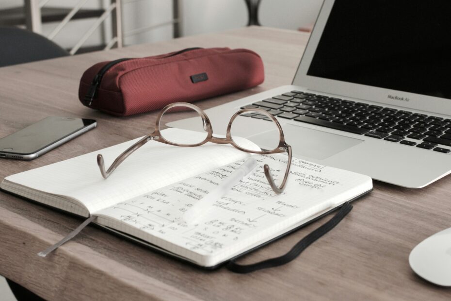 A work desk with a laptop, a notebook filled with handwritten notes, and a pair of eyeglasses resting on the open pages. There is also a pen placed on the notebook, a smartphone to the left, and a red pencil case behind the notebook. The setup suggests a workspace for studying or working, with a focus on writing or note-taking.