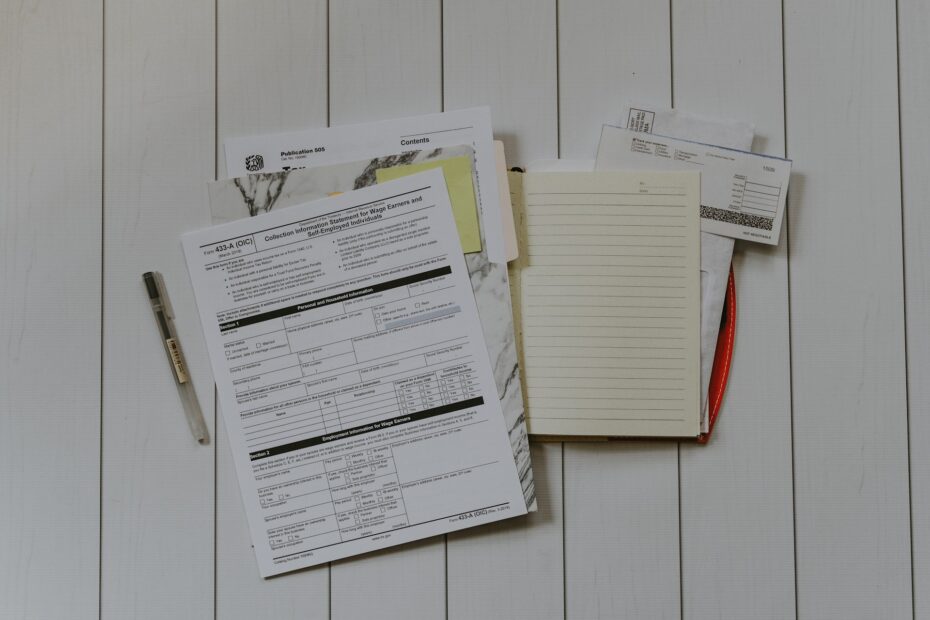 A pen and several documents, including tax forms and other paperwork, are laid out on a white wooden surface next to an open, lined notebook.