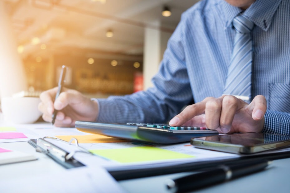 A person in a business setting is working with financial documents, using a calculator, and holding a pen. The individual appears focused, with a smartphone and clipboard on the desk, along with colorful sticky notes. Sunlight softly illuminates the workspace, adding a professional and organized atmosphere.