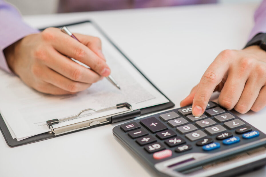 A person is calculating finances using a calculator and taking notes on a clipboard, possibly reviewing financial statements or preparing a budget.