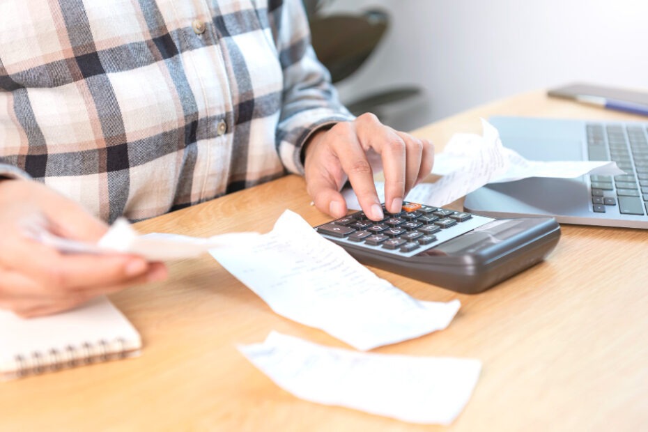 A person calculating finances using a calculator with receipts and a laptop on a desk.