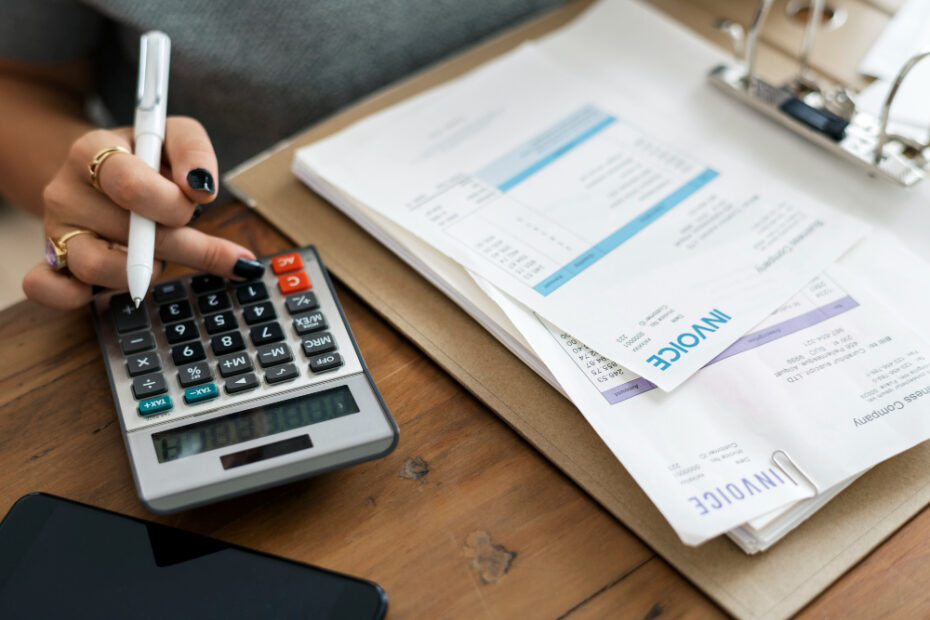 A close-up image showing a person using a calculator while holding a pen, with several invoices spread out on a clipboard. The scene represents the process of managing finances, calculating expenses, and organizing bills, emphasizing the importance of accurate invoicing and financial documentation.