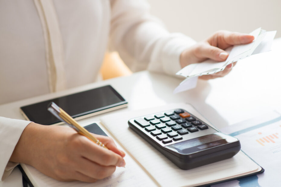 A person is sitting at a desk with a notepad, calculator, and smartphone. They are holding a receipt or invoice in one hand while writing notes with the other hand. The calculator and the phone are placed on the desk, and the environment suggests someone working on financial tasks, such as budgeting or accounting.