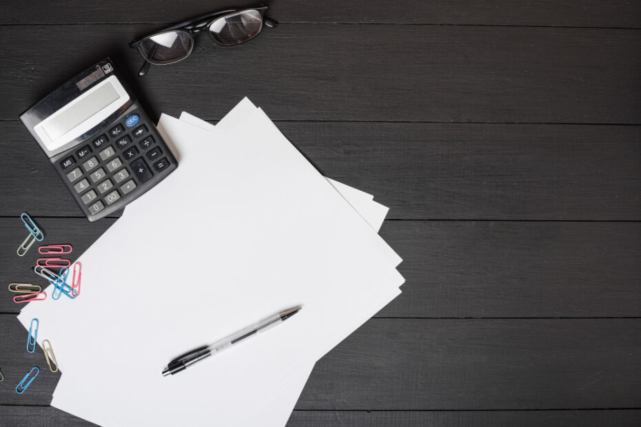 A neat and organized workspace setup on a dark wooden table featuring a calculator, several sheets of blank white paper, a pen, and paper clips scattered around. The calculator and a pair of glasses are placed to the left, suggesting that financial calculations or administrative work might be underway.