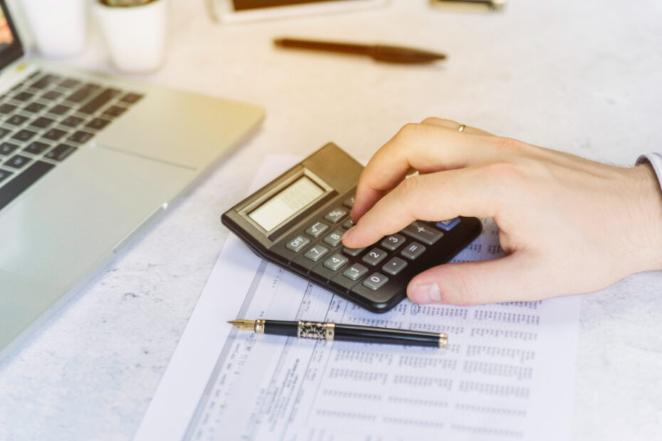 A person is using a calculator to work on financial calculations, with a laptop, a pen, and some documents visible on the desk. This image captures a moment of financial management or accounting tasks being performed in an office setting.