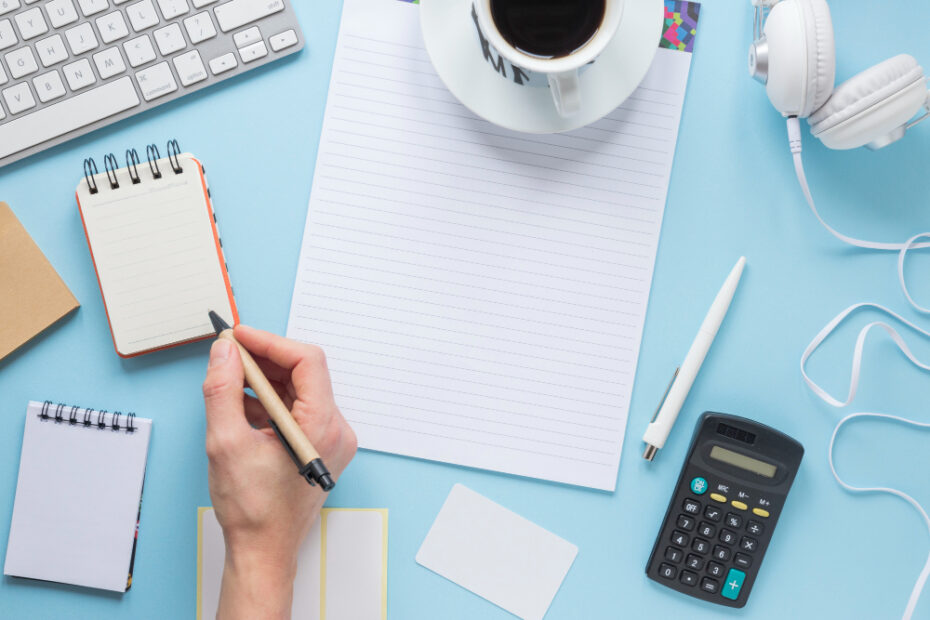 A workspace setup featuring a person's hand holding a pen, ready to write on a small notepad. The desk includes a blank sheet of paper, a cup of coffee, a calculator, headphones, and a keyboard, all neatly arranged on a light blue surface, conveying an organized and productive environment.