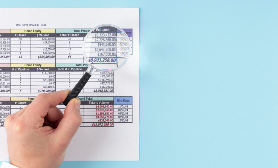 The image shows a hand holding a magnifying glass, zooming in on a financial document or report. The report contains multiple tables with columns listing details like "Home Equity," "Total Production," and other financial metrics such as "$ Volume," "# Closed," and totals. It also includes a column for "Hire Date," indicating dates associated with employee records or performance. The magnifying glass highlights some of the larger figures in the report, suggesting a focus on key financial data, possibly for analysis or review purposes.