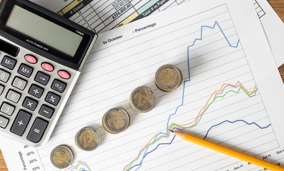 This image displays a financial analysis setup with a calculator, a pencil, and some coins placed on sheets of paper with printed graphs. The graphs show trends and data points in different colors, indicating fluctuations over time, likely representing changes in financial metrics such as sales, revenue, or stock performance.