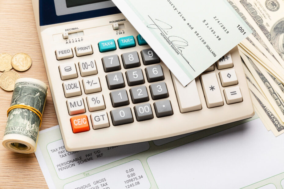 A detailed image of a financial workspace setup. It features a beige calculator prominently displayed with various function keys visible, such as tax percentage and grand total. Beside the calculator, there are rolled up U.S. dollar bills, loose coins, and a check resting on a stack of paper currency. The check shows details typical of payroll or business transactions, including signatures and monetary amounts. The setup suggests financial calculations, possibly for accounting or tax preparation purposes