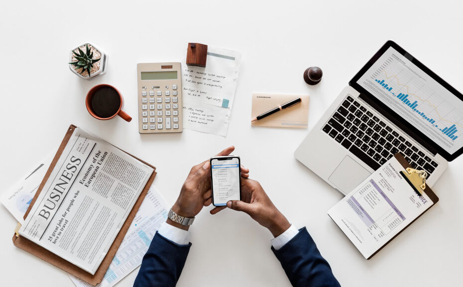 A person is sitting at a desk surrounded by business-related items. On the desk, there's a laptop displaying financial graphs, a calculator, a clipboard with documents, a newspaper with business news, and a cup of coffee. The person is holding a smartphone, presumably checking or entering data. This setup suggests a professional environment where financial or business activities are being managed.