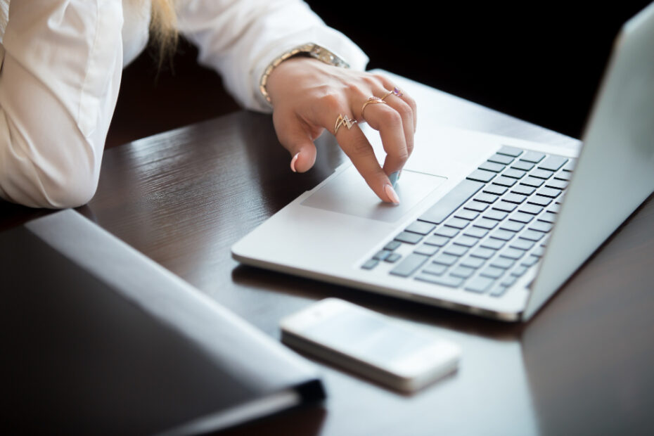 A woman in a white blouse is working on a laptop in a business environment. Her focus is on the laptop's trackpad as she navigates through tasks, with her smartphone positioned nearby on the table, suggesting a professional setting and multi-tasking scenario.