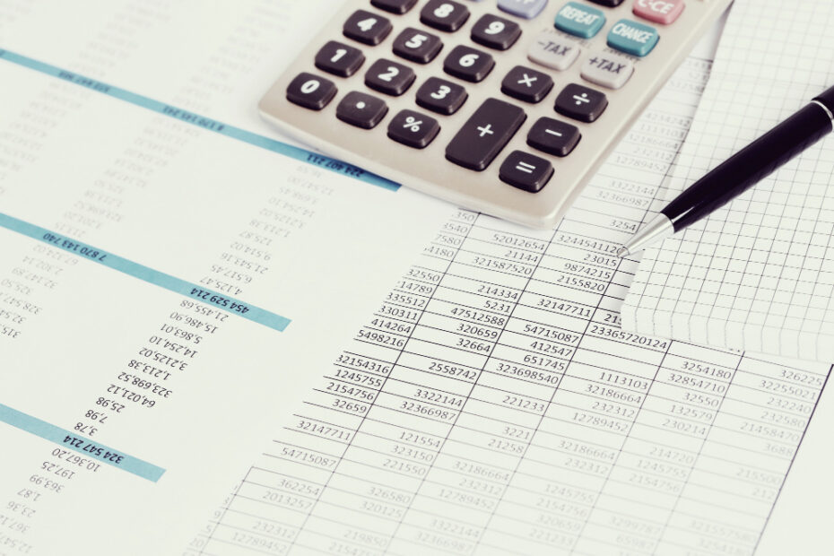 A close-up image of financial documents showing tables with numbers, a calculator, and a pen resting on a graph paper notebook. The scene represents financial calculations, accounting, or data analysis typically used in creating invoices, reports, or balance sheets.