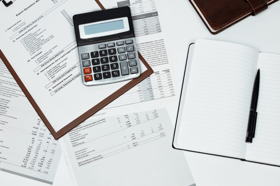 A calculator, financial documents, and an open notebook with a pen are laid out on a desk, indicating a workspace focused on financial analysis or accounting.