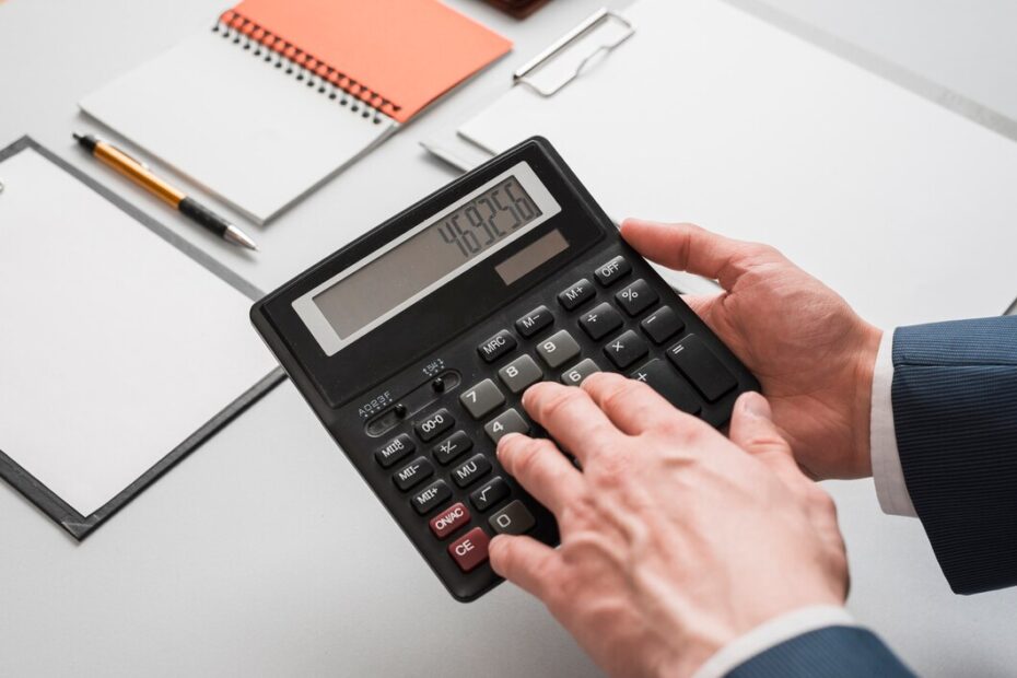 A person calculating numbers on a calculator with notepad and pens in the background on a desk.