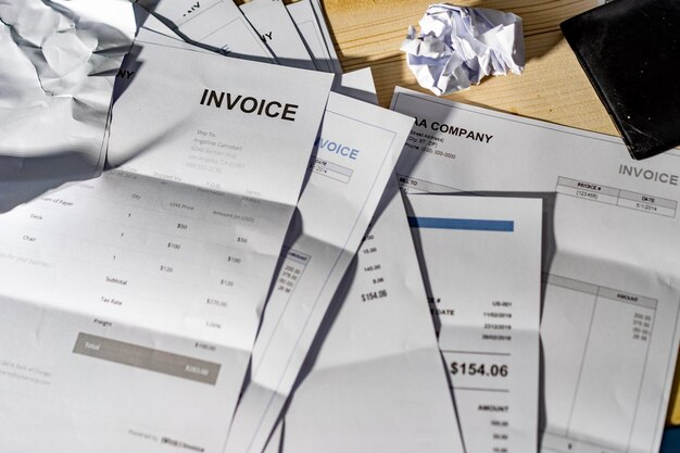 A scattered collection of invoices, some crumpled, lying on a wooden desk. The invoices feature different amounts and company details, with one prominently displaying the word "INVOICE" at the top. The scene suggests a busy or chaotic environment, possibly indicative of financial paperwork being organized or reviewed.