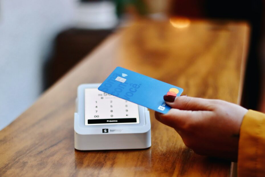 The image depicts a person making a payment using a credit card at a modern card payment terminal. The terminal is small and portable, sitting on a wooden counter, indicating a contemporary and convenient method for financial transactions