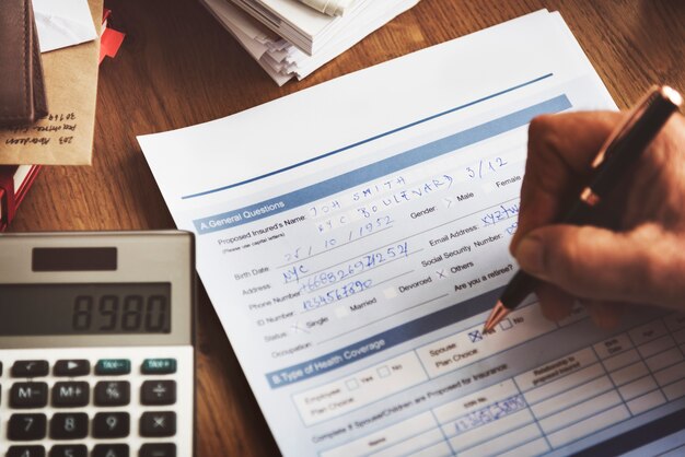 A close-up image showing a person filling out a form with a pen. The form appears to be related to personal or health information, with fields for name, address, date of birth, and other details. A calculator is positioned nearby, along with a stack of documents, indicating a task involving data entry or financial calculations. The scene suggests a work environment focused on detailed paperwork or administrative tasks.