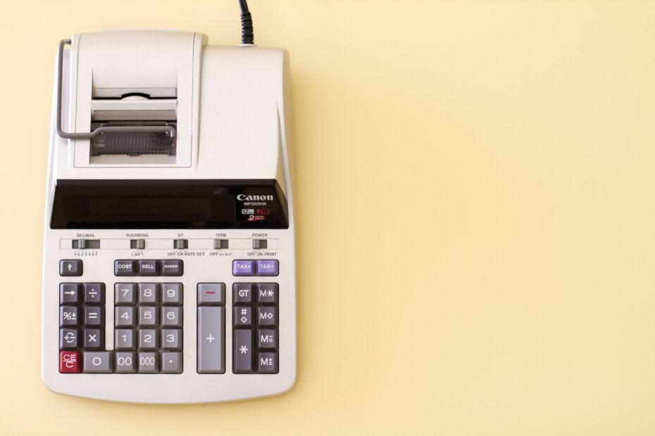 An image of a Canon MP20DHIII calculator. This desktop calculator features a printing roll at the top, and a keypad with large, clearly marked buttons for various functions including cost, sell, and margin calculations, as well as settings for tax, grand total, and item count. The background is a plain light yellow, emphasizing the calculator's presence.