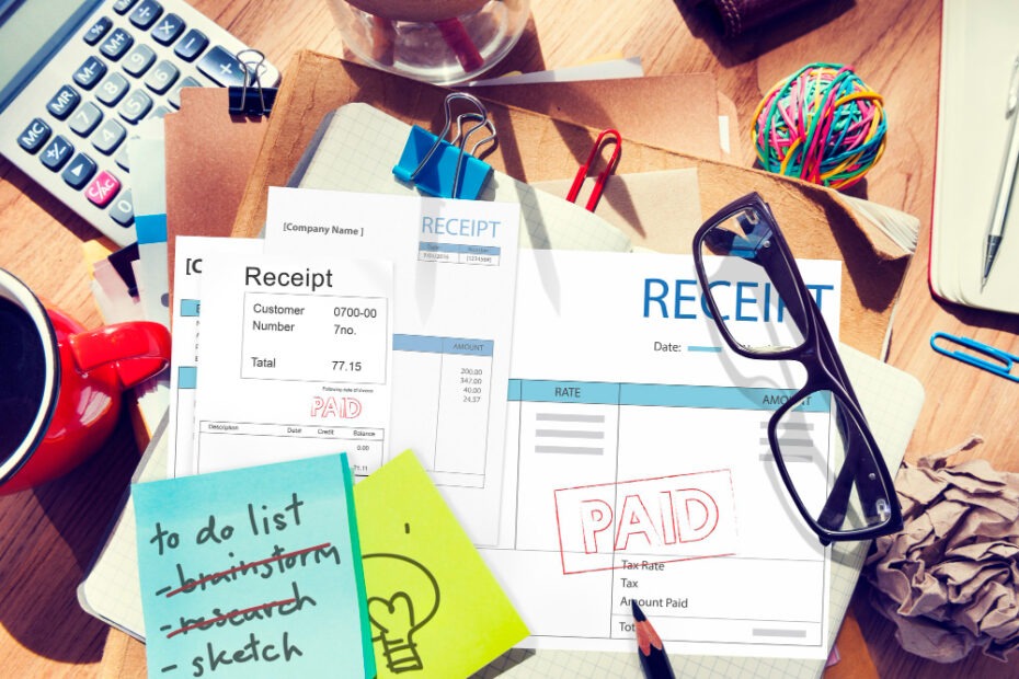 An overhead view of a cluttered desk featuring various office items, including receipts marked "PAID," glasses, a cup of coffee, a calculator, colorful rubber bands, and sticky notes. One sticky note has a "to-do list" with tasks such as "brainstorm" and "research" crossed off, leaving "sketch" as the remaining task. The scene reflects a busy workspace focused on managing finances and completing tasks.